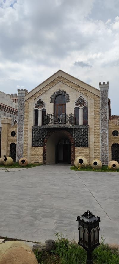 The image shows the exterior of a stone building, likely a winery, in Voskevaz, Armenia. The building has a grand entrance with an arched doorway, ornate balconies, and decorative stonework. Large clay amphorae are placed around the entrance, suggesting a connection to wine production. A paved courtyard leads to the entrance, and the sky above is overcast. There are no people visible in the image. The architectural style appears to be a blend of traditional Armenian elements with more decorative, perhaps gothic-inspired, features. The overall impression is one of historical significance and artisanal craft.