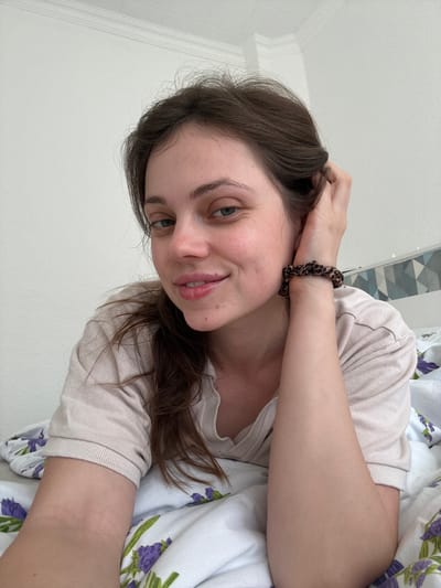 A woman with brown hair and blue eyes is lying in bed. She is wearing a light-colored shirt and a leopard print scrunchie on her wrist. The bed is covered with white bedding with a floral pattern. The background includes a white wall and a decorative tiled wall. The photograph does not include any location of Kőröshegy, Hungary.