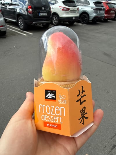 The image displays a fair-skinned hand holding a frozen dessert in an outdoor parking lot. The dessert itself is a colorful frozen treat, visible through a clear plastic container, exhibiting a gradient from red/pink at the top to yellow/orange at the bottom, resembling a mango. This container is partially encased in an orange cardboard sleeve.
The cardboard sleeve features several printed details. The brand logo "FRIENDS OF Asia" is visible, along with a "VEGAN" certification symbol. The English text "frozen dessert" is printed vertically, and "MANGO" clearly indicates the flavor. Above this, two Asian characters, likely Chinese for "mango" (芒果), are written in black.
In the background, several cars are parked on an asphalt surface, characteristic of a parking lot. The vehicles include a white SUV, a red car, and a dark grey SUV or crossover. A partial license plate "S B 809" and a "Duster" badge are discernible on the dark grey car, confirming its model as a Dacia Duster and its German origin. The scene is brightly lit, indicating daytime. The overall setting is an ordinary outdoor environment, consistent with the provided location context of Waldshut, Germany.