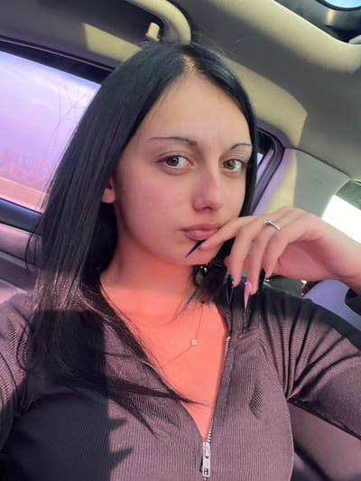 A female individual is depicted in a close-up portrait taken within a vehicle interior, located in Akhalsopeli, Georgia. She possesses long, straight black hair parted down the center, dark brown eyes, thin arched eyebrows, and full lips. She is dressed in a dark, ribbed top featuring a silver zipper closure down the front. A gold-colored chain necklace with a small, white four-leaf clover pendant is visible on her neck. On the ring finger of her right hand, a silver-colored ring with a clear stone is present. Her right hand is raised and positioned near her chin, with fingers slightly curled. The fingernails on this hand are notably long, pointed, and painted in a dark color, transitioning to a lighter pink hue near the base on some digits. The vehicle's interior is visible with a light-colored headliner and a blurred side window displaying a reddish-purple ambient light from an outdoor scene. Strong, warm directional lighting illuminates the subject from her right side, casting a reddish glow on her skin and clothing.