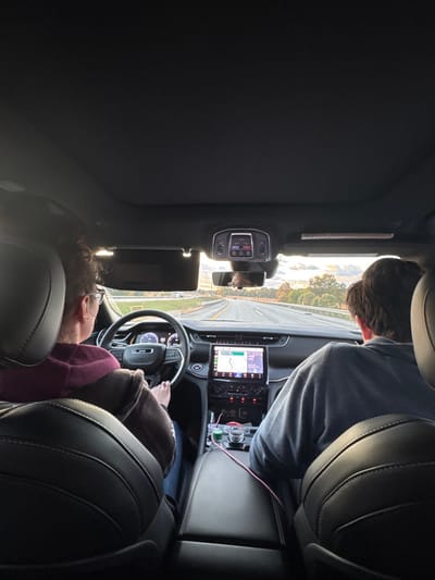 This image captures the interior of a moving Jeep vehicle, observed from a rear passenger seat, looking forward through the windshield. The vehicle is traveling on a multi-lane asphalt highway, reportedly in or near South Bend, United States. The surrounding environment consists of metal guardrails lining the highway, with trees displaying autumnal foliage in the distance under a partially cloudy sky featuring warm hues near the horizon, indicative of late afternoon or early morning.
The driver, an adult female with short, dark, curly hair and glasses, wearing a maroon hooded sweatshirt, is seated behind the multi-function steering wheel, actively operating the vehicle. Her hands are positioned on the steering wheel. The digital instrument cluster behind the steering wheel displays a speed of approximately 70-75 mph.
In the front passenger seat, an adult male or young adult with brown, medium-length hair, wearing a grey long-sleeved shirt, is seated. The vehicle's interior features black leather seats and a black dashboard. A prominent vertical infotainment touchscreen in the center console displays a navigation map. Below this screen, a rotary gear selector and other climate/media controls are visible. A pink charging cable and a green cable are plugged into ports within the center console area. The rearview mirror shows a partial, blurred reflection.