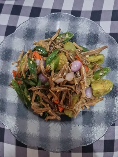 A plate of Indonesian cuisine, Teri Balado, sits on a checkered surface. The dish is a mix of fried anchovies (teri), green tomatoes, green chili peppers, shallots, and red chili peppers. The ingredients are combined and arranged in a haphazard way in a glass dish. The chili peppers are cut into small pieces, and the tomatoes are quartered. The shallots are small and round.