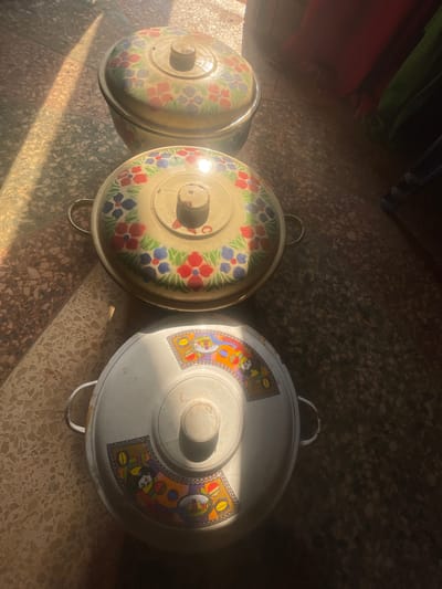 The scene depicts three circular, lidded food warmers or serving dishes arranged diagonally on a tiled floor in Awgbu, Nigeria. The dishes are positioned from the lower-right foreground to the upper-left background. The bottom-most and largest dish is white enamelware, featuring a lid decorated with radial segments containing colorful cartoon characters, including a clown, and possibly text, around a central white knob. It has two small, metallic loop handles. The middle dish, slightly smaller, is an off-white or light yellow enamelware with a lid and sides adorned with a repeating floral pattern of red and blue flowers with green leaves. It also has a central knob and two small, metallic loop handles. The top-most dish is the smallest, similar in color and floral pattern to the middle dish, with a central knob on its lid. All three items exhibit signs of use and minor wear. The floor is composed of light-colored, speckled tiles. Strong sunlight streams from the left, casting bright bands and elongated shadows across the floor and illuminating parts of the dishes. The background on the right is blurred and darker, showing indistinct shapes and colors, possibly fabrics or household items. No people are visible.
