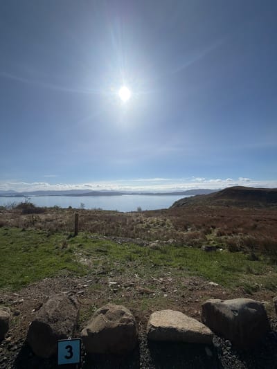The image shows a landscape in Portree, United Kingdom, with a bright sun illuminating the sky on the left. A body of water, likely a loch or bay, stretches horizontally across the middle of the frame, separating the sky from a landmass. The landmass on the right side of the image consists of grassy terrain with brown, dry patches, suggesting late winter or early spring. Several large rocks are positioned near the bottom right corner. A small sign with the number "3" is visible near the rocks. The scene suggests a hiking trail overlooking the water.