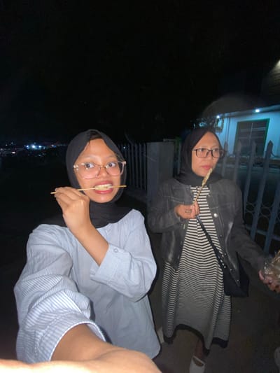 Two young women wearing hijabs are standing outside at night. The woman in the foreground is wearing a light blue and white striped shirt and has a skewer of food in her mouth. She is wearing glasses. The woman behind her is wearing a black denim jacket over a black and white striped dress and also wearing glasses. She is holding a skewer of food. The background shows a dimly lit residential area with houses and fences. The scene appears to be a street or pathway in a neighborhood, potentially in the city of Tanggul, Indonesia, given the context.