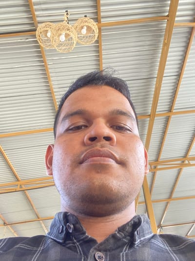 A low-angle, close-up selfie shows a person's face and upper torso. The person has dark hair, brown eyes, and a neutral expression. They are wearing a dark, plaid button-up shirt. The background consists of a corrugated metal ceiling with wooden beams and three woven pendant light fixtures. The lighting suggests an indoor or covered outdoor location. The provided information states this scene is located in Aluepiet, Indonesia.