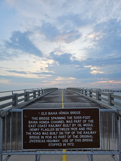 The image shows the Old Bahia Honda Bridge in Big Pine Key, United States. The bridge stretches into the distance under a cloudy sky. A sign attached to the bridge railing provides historical information: the bridge spans 5055 feet across the Bahia Honda Channel and was originally part of the East Coast Railway, built by Henry Flagler between 1905 and 1912. The road was built on top of the railway bridge in 1938 as part of the original Overseas Highway. The bridge was no longer in use after 1972. A yellow line is visible along the surface of the bridge. The railings are constructed of metal and concrete.