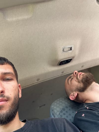 The image captures two individuals inside a vehicle, likely a van, suggesting an indoor setting. One individual is positioned in the foreground, looking directly at the camera, with their face partially visible from the nose upwards. They have short, dark hair and a beard. The second individual is in the background, lying down with their head resting on a patterned seat. This person has a beard and is wearing a dark sweatshirt with a pattern of pink flamingos. Above them, the vehicle's ceiling is visible, featuring a light fixture and climate control knobs. The lighting suggests it's daytime. The image does not provide any specific visual information to pinpoint the location within Tulovo, Bulgaria.