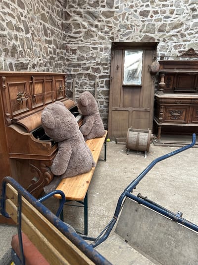 The image depicts an indoor scene with a stone wall as the backdrop. In the foreground, a vintage wooden piano sits open, with two large, plush teddy bears seated on a wooden bench in front of it, appearing to play the instrument. Behind the piano, a tall wooden cabinet with a mirror and coat hooks stands against the wall. To the right of the cabinet are two ornate wooden furniture pieces, likely antique dressers or sideboards. In the middle ground, a small, wooden barrel-like object rests on the concrete floor. A blue metal cart with wooden components is partially visible in the lower right corner. The overall setting suggests a storage area or an antique shop. The image location is Gonneville-la-Mallet, France.