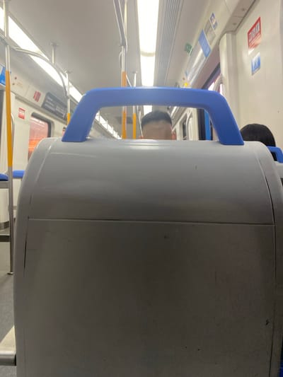 The image displays the interior of a passenger train car, presumably on a line operating within or near Luis Guillón, Argentina. In the foreground, the back of a light gray padded train seat is visible, featuring a prominent blue plastic grab handle mounted horizontally on its upper edge. Partially obscured by this seat, the upper portion of a person's head, characterized by short, dark hair and a visible forehead, is seen. Further back and to the right, a second individual's dark-haired head is visible, partially obscured. The train's interior features white walls and ceiling, equipped with multiple fluorescent light fixtures running lengthwise. Silver-colored horizontal and vertical grab bars are present, along with bright yellow vertical support poles. Windows along the left side of the frame show an indistinct reddish-orange glow from the exterior, suggesting ambient light or an external reflection. Various rectangular signs are affixed to the train car's walls, some displaying unreadable text or pictograms; one sign on the left wall shows the numbers "500.000." No specific actions or interactions are depicted; the scene captures a static moment within a moving or stationary public transport vehicle.