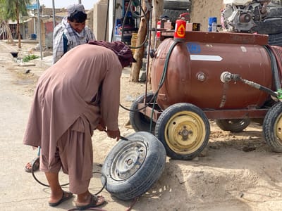 The image shows two men in Panjwai, Afghanistan, at coordinates 31.542, 65.463. The man on the left, wearing a white and brown patterned long-sleeved shirt, stands near a tire of a small trailer with an air compressor tank. The tank is painted brown with a white stripe, and has hoses connected to it. The second man, wearing a brown long tunic, is on his knees, inserting a tire inflator hose into a tire. The tire appears to be on a trailer, which is attached to the air compressor tank. The tank is mounted on a steel frame, and has a small engine and spare tires behind it. The scene is set on a dusty, dirt road in Panjwai.