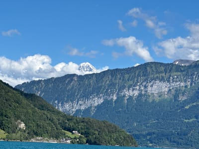 This image shows a panoramic view of Lake Thun, Switzerland. The foreground features the bright turquoise waters of the lake, with gentle ripples on its surface. The middle ground is dominated by steep, tree-covered mountains that slope down to the water's edge. Several small houses and buildings are nestled amongst the trees and on the lower slopes, suggesting a scattered settlement. In the background, majestic, snow-capped mountains rise sharply against a clear blue sky dotted with white, puffy clouds. The lighting suggests it is daytime, likely mid-morning or afternoon, with bright sunshine illuminating the scene.