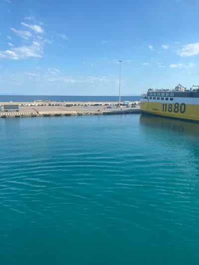 In the Municipal Unit of Kastro - Kyllini, Greece, a large ferry, predominantly yellow and white, is docked at a port. The ferry's hull displays the number "11880" in large black numerals. The port area features a parking lot with several cars and a truck, a concrete barrier, and a tall lamppost. The bright blue sky is dotted with scattered white clouds. The foreground is dominated by the clear, turquoise water of the sea, with gentle ripples on its surface. The sea extends to the horizon, where distant landmasses are visible. A group of people are visible on the deck of the ferry, suggesting activity and movement.