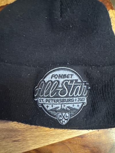 A black knit beanie rests on a wooden surface. The beanie features a prominent circular embroidered logo rendered in light grey and silver threads. The logo design includes the text "FONIBET" at the top, "All-Star" in a stylized script centrally, and "ST. PETERSBURG + 2023" along the lower arc. Below the "All-Star" text, a detailed emblem with wings, stars, and a shield containing the letters "KHL" is visible. The black fabric of the beanie shows minor lint accumulation. The underlying wooden surface displays distinct grain patterns. This scene is situated in Matveevskoe, Russia.