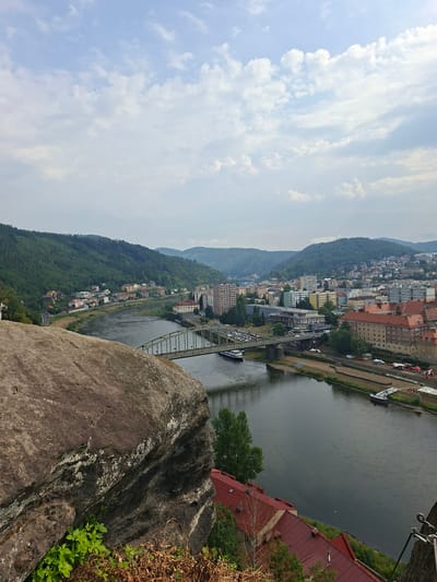 This is a wide-angle, eye-level shot of the Elbe River flowing through Děčín, Czechia. In the foreground, a large, weathered rock formation occupies the lower left quadrant. Beyond the rock, the Elbe River stretches across the frame, its dark water reflecting the overcast sky. A bridge with a distinctive green metal structure spans the river in the middle ground, with a small white boat passing beneath it.

The left bank of the river features a densely wooded hill, receding into the distance. Along the base of the hill, a small town with colorful houses is visible, nestled by the river's edge. The right bank of the river is lined with a more urban landscape. Buildings of varying heights and architectural styles, including apartment blocks and older brick structures with red-tiled roofs, dominate this side of the city. The cityscape extends up the gentle slope of the hills in the background, which are also covered in a mix of trees and residential areas. The sky is a pale blue, with scattered white clouds. A few trees and some dried vegetation are visible at the bottom edge of the frame.