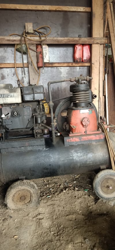 The image depicts an old, possibly disused, air compressor unit situated on a dirt floor in what appears to be a workshop or storage area. The compressor has a black, cylindrical tank and a red and black compressor head with fins. A Kubota engine is attached to the side of the compressor. Various hoses and pipes are connected to the unit. The background shows wooden beams and a gray metal wall. Two red plastic containers are visible on a shelf above the compressor. The ground around the compressor is littered with debris, including what appear to be cigarette butts. The overall impression is one of a functional but neglected piece of machinery in an outdoor or semi-outdoor setting. The location in Uteungeulinggang, Indonesia, suggests a tropical or subtropical environment, which might explain the presence of dirt and debris. There are no people or animals visible in the image.