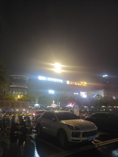 This image depicts a street scene at night in Nanhai District, China, likely after rain, as evidenced by wet surfaces and reflections. In the foreground, a white Porsche Cayenne is parked, its license plate clearly visible as "A-7F76B". To its right, a dark-colored sedan is also parked. Several motorcycles are parked in a designated area behind a metal fence.
A group of individuals, some holding umbrellas, are visible near the motorcycles. The background is dominated by a multi-story building with illuminated signage. Prominent signs include "LIVE HOUSE" in large orange letters, and other Chinese characters that suggest commercial establishments such as " OASIS BILLIARDS CLUB" and "美团", a food delivery service. The lighting from the buildings and streetlights creates a vibrant but somewhat hazy atmosphere. The sky is dark, indicating nighttime. The overall scene suggests a busy urban environment with commercial activity.