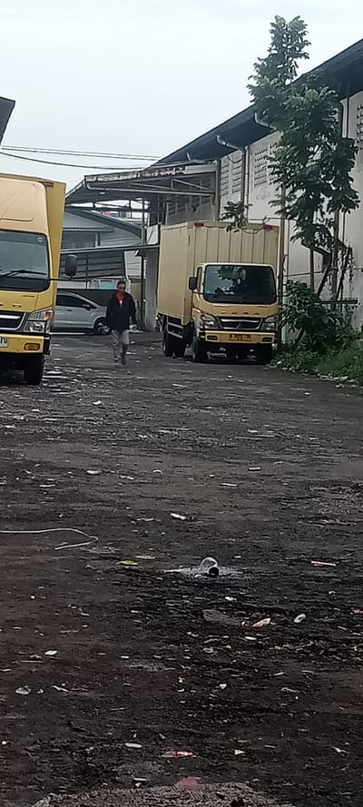 The image depicts an outdoor industrial or commercial area in Sulaeman, Indonesia, under an overcast sky. The foreground and mid-ground consist of an unpaved, dark, uneven dirt and gravel surface, heavily littered with various debris including plastic items, paper, and what appears to be a coiled white hose or cable, and a crushed metal or plastic can. Puddles are visible on the ground.

One male individual is positioned in the mid-ground, walking forward towards the viewer, slightly to the right of the image center. He wears a dark jacket over a red-collared shirt, light-colored pants, and dark shoes. He has glasses or sunglasses and his head is slightly tilted downwards.

To the left, the front portion of a yellow box truck is partially visible, showing a license plate fragment "TU". In the mid-ground, behind the individual and to the right, another yellow box truck is parked, facing the camera. Its license plate, "N 9226 TX," is clearly visible. Further in the background, between the individual and the left truck, a white sedan is partially discernible.

Structures include a large white building on the right with a dark sloped roof, a white downspout, and multiple horizontal vents or window slats. A small tree with green foliage grows near this building. In the background, between the trucks, a darker, possibly metal-roofed structure with support beams is visible. Overhead power lines run horizontally across the upper-mid section of the image.