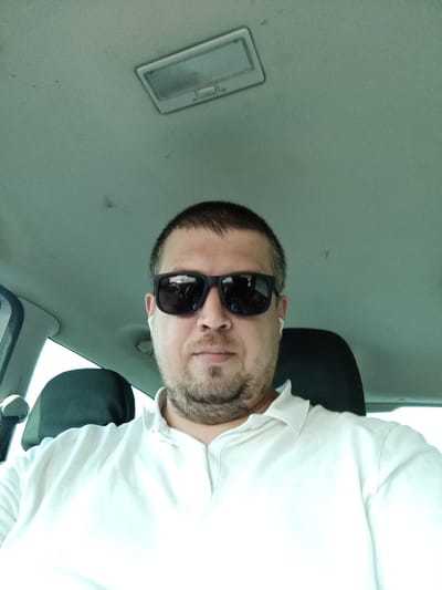 A man wearing sunglasses and a white polo shirt is sitting in the driver's seat of a car. He has short dark hair and a beard. The interior of the car is visible, including the ceiling light and the headrests of the seats. The lighting suggests it is daytime. The image was taken in Třemošnice, Czechia.