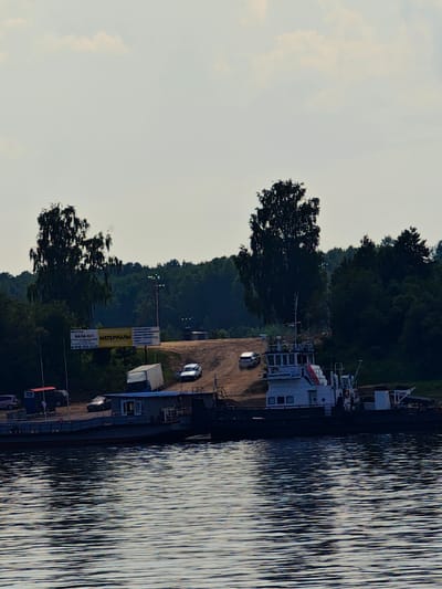 The image depicts a waterfront scene in Erkalovo, Russia. A tugboat is moored to a ferry, which is partially submerged in the water. The ferry is loaded with what appears to be building materials, including a white truck and a blue modular building. In the background, a dirt ramp leads up to a wooded area, and several vehicles are parked near a sign advertising "MATERIALS". The sky is overcast, with scattered clouds. The overall atmosphere is one of industrial activity and transportation.