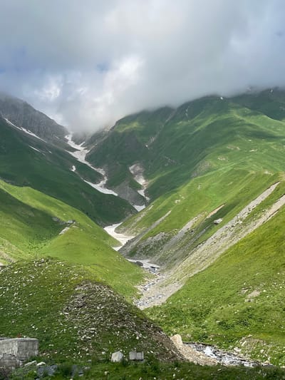 The image depicts a lush green mountain valley, with steep slopes covered in vibrant grass. Patches of snow are visible in the upper parts of the valley, indicating a high altitude. A winding stream flows through the valley floor, bordered by rocks and gravel. The sky is partly cloudy, with some of the clouds obscuring the mountain peaks. In the foreground, there are remnants of man-made structures, appearing to be concrete foundations or walls, with some scattered stones and vegetation. The scene conveys a sense of natural beauty and remote wilderness, characteristic of mountainous regions. This scenery is consistent with the landscape found in the vicinity of Almasiani, Georgia, known for its dramatic mountain ranges.