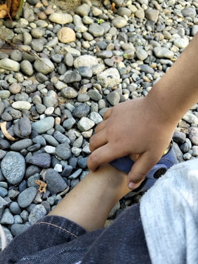 Location: Panton, Indonesia.
A high-angle, close-up photograph depicts a child's right hand interacting with their left leg over a ground surface composed entirely of small, rounded pebbles. The child's skin tone is light brown. The right hand, extending from the upper right, appears to be touching the child's left ankle, where a portion of a blue sandal or shoe is visible. The left leg is partially covered by dark blue denim fabric with orange stitching, consistent with jeans, and a light grey garment is visible on the far right. The ground is densely covered with smooth, multi-colored pebbles of varying sizes, predominantly grey, brown, and light blue, with sparse dried leaf litter.