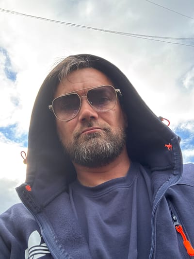 A medium shot, taken from a low angle, shows a man wearing a dark blue hooded jacket and a dark blue t-shirt. He has short, brown hair with some grey mixed in, a beard, and is wearing aviator-style sunglasses with a gold frame. The hood of his jacket is up, partially obscuring the top of his head and framing his face. The background consists of a cloudy sky with some blue patches and power lines running horizontally across the top of the frame. The man is looking directly at the camera. The scene appears to be outdoors in Usinsk, Russia, given the cloudy sky and the context.