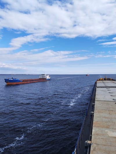 The image depicts a waterway near Maksakovo, Russia. The scene is viewed from the deck of a barge. Another blue and orange barge is moving away in the distance, its name clearly visible as "OKSINIYA" on its side. Further away, a third barge is visible. The water is dark blue with white wakes visible behind the barges. The sky is bright blue with scattered white clouds.