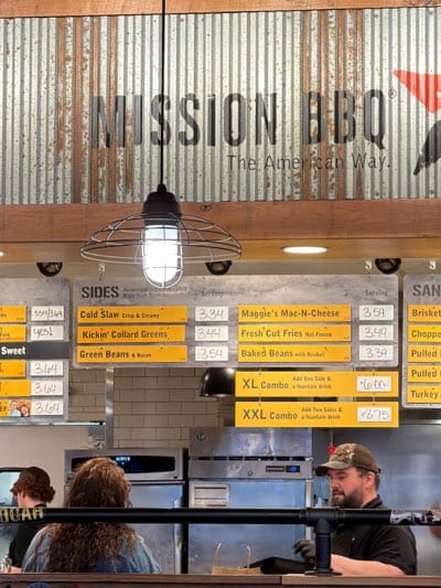 The image shows the interior of a Mission BBQ restaurant, likely located in Rochester, United States, based on the assumption the image was taken there. The "MISSION BBQ - The American Way" sign is visible at the top of the frame, mounted on a corrugated metal wall with wooden trim. Below the sign is a menu board listing sides, including "Cold Slaw," "Kickin' Collard Greens," "Green Beans & Bacon," "Maggie's Mac-N-Cheese," "Fresh Cut Fries," and "Baked Beans." The prices are listed beside each item. Combination meals, "XL Combo" and "XXL Combo," are also listed with their prices. To the right, a "SAND" menu is partially visible, with "Brisket," "Chopped," "Pulled," and "Turkey" listed.
Below the menu, there's a counter where customers are being served. A person with a camo hat and a beard, likely an employee, is visible behind the counter. Two customers are in front of the counter, partially seen from behind. Various pieces of restaurant equipment are visible in the background behind the employee. An industrial-style light fixture hangs from the ceiling.