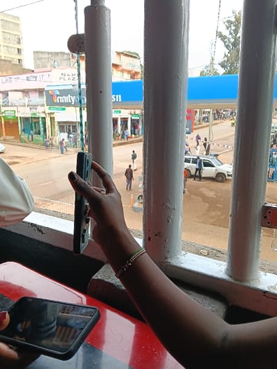 The image captures a street scene viewed from indoors, looking out through a window or barrier framed by white vertical bars. In the immediate foreground, a person's dark-skinned arm is prominent on the right, holding a black smartphone vertically with the screen facing away from the viewer. A multi-colored beaded bracelet, featuring green, white, and red beads, is worn on the wrist. Another black smartphone is visible lying horizontally on a red and black table surface in the bottom left, reflecting the surroundings. Part of a white sleeve or garment is also visible on the far left. Between two of the white vertical bars, a dark green cord with a small grey handle or weight hangs down, possibly a pull for a blind or light.
The street scene outside reveals an urban or semi-urban environment. The road appears to be made of dirt or is unpaved in some sections, with light brown tones. Several people are walking on the sidewalks and street. On the right side, a petrol station with a large blue canopy is visible, bearing the letters "sII" (likely part of "Shell" or a similar brand name). A red sign at one of the petrol pumps displays "30.00." A white SUV-like vehicle is parked or moving slowly near the petrol station, with a person in a light blue shirt standing nearby.
On the left side of the street, various buildings with storefronts line the road. Visible text on the buildings includes "NAMI PLAZA" and "Trinity Plaza" on a multi-story building, and further left, partially obscured text reads "LIKE IT..." on a wall. The sky is overcast, indicating daytime but without direct sunlight. The overall impression is a bustling street view, possibly in Kenya, given the colors of the bracelet resembling the Kenyan flag.