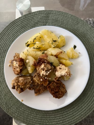 The image shows a white plate of food placed on a green, woven placemat. The plate contains boiled potatoes seasoned with dill and what appear to be pieces of fried meat or cutlets with a creamy filling. The food is arranged on the plate, with the potatoes towards the top and the meat dishes towards the bottom and left. The placemat is round and has a textured pattern. In the background, a clear glass surface of a table is visible, with a metallic cylindrical object on it, possibly a leg or support for the table. The overall setting suggests a meal being served on a table, likely indoors. There is no information in the image that could indicate a specific location in the city of Olomouc, Czechia.