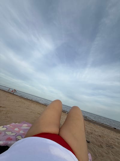 The image depicts a first-person perspective of someone relaxing on a sandy beach, with the ocean and a cloudy sky in the background. The foreground shows the person's legs, clad in red swimwear, and a white garment. A towel with a heart pattern is visible underneath. In the mid-ground, a few people are visible in the shallow water. The location is identified as the city of Ondvor, Russia.
