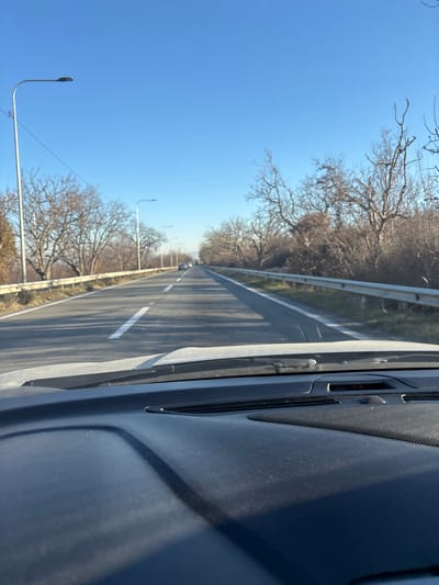 The image displays a two-lane paved road in Changilari, Georgia, viewed from the interior of a vehicle. The foreground exhibits a dark, dust-covered dashboard and the lower portion of the windshield, including a wiper blade. The road features white lane markings, with a broken centerline and a solid white line defining the right edge. Metal guardrails run along both sides of the road. Beyond these guardrails, numerous bare deciduous trees and dense brush line the path, suggesting a dormant season. Multiple streetlights with dark, elongated fixtures are spaced along the left side of the road, receding into the distance. Several vehicles are visible traveling in the same direction in the far distance. A clear, bright blue sky occupies the upper frame, with a faint, hazy outline of distant landforms visible on the horizon. The scene is illuminated by direct sunlight.