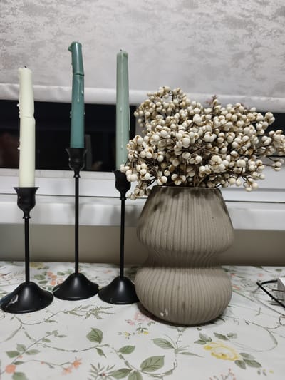 A still life arrangement is set on a floral-patterned tablecloth, featuring three tall, slender candles in black candleholders of varying heights. The candles themselves are of different colors and thicknesses; the leftmost is a pale cream and appears to have some melting, the middle one is a deep teal and is significantly melted at the top, and the rightmost is a lighter, muted green and also shows signs of melting. To the right of the candleholders is a textured, beige vase with a ribbed surface, holding a dense bouquet of small, round, white berries on thin, brown stems. The background consists of a textured, off-white surface, possibly a wall or window covering, with horizontal lines suggesting blinds or a screen. The overall scene is illuminated with soft, artificial light. There are no people visible in the image. The location is not specified in the image or the provided crops.