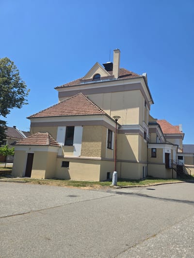 The image depicts a modern building with a red-tiled roof, situated next to an asphalt road. The building has a light yellow facade with beige and brown decorative elements. Several windows of varying sizes are visible, some with dark frames. A metal staircase leads to an upper-level entrance on one side of the building. In front of the building, a small patch of grass borders the road, and a grey cylindrical object, possibly a utility box or post, stands near the edge of the asphalt. The sky is clear and blue, indicating a sunny day. The scene appears to be a residential or commercial area in a town. The image is oriented vertically, as if taken from a slightly elevated perspective looking down. No people are visible in the image, and no text is discernible. The lighting suggests it is daytime, likely late morning or early afternoon due to the clear, bright sunlight and distinct shadows cast by the building.