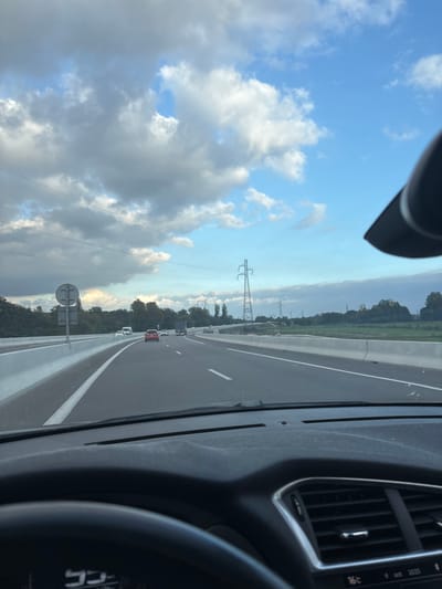 The image presents a forensic view from the driver's perspective inside a vehicle traveling on a multi-lane highway in Châtenois, France. In the immediate foreground, the dark dashboard of the vehicle is visible, along with a portion of the steering wheel on the lower left and air vents on the right. A digital display on the right side of the dashboard shows "16°C" and "oct 2025." A fine layer of dust is present on the dashboard surface.
Ahead, the highway extends with at least two lanes of travel, demarcated by solid white lines. Concrete barriers line both the left median and the right shoulder of the road. A partially obscured circular road sign is mounted on the left median barrier. Multiple vehicles are in motion ahead in the same direction, including a red sedan in the middle lane, a white van further ahead in the left lane, and a dark truck in the right lane.
The environment adjacent to the highway features dense green foliage and trees along both sides. A tall metal lattice electricity transmission tower stands prominently in the middle distance on the right side of the road, with power lines extending across the sky. The sky above is partly cloudy, displaying significant white cumulus clouds against patches of clear blue, under lighting consistent with late afternoon or early evening.