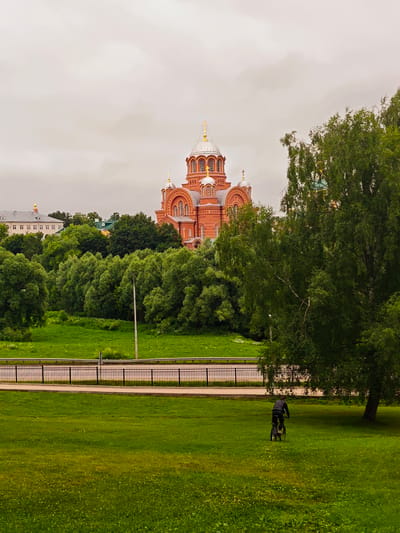 The image shows a landscape view of the city of Khotkovo, Russia, featuring the Holy Trinity St. Sergius Lavra's bell tower and the surrounding green space. In the foreground, a person is riding a bicycle on a grassy field. A road with a black metal fence runs horizontally across the middle ground, separating the field from a densely wooded area. Behind the trees, the prominent red brick structure of the Holy Trinity St. Sergius Lavra, with its silver domes and golden crosses, is visible against an overcast sky. Further in the background, another building with a white facade and a golden dome can be seen. The overall scene is dominated by lush greenery and a cloudy sky.