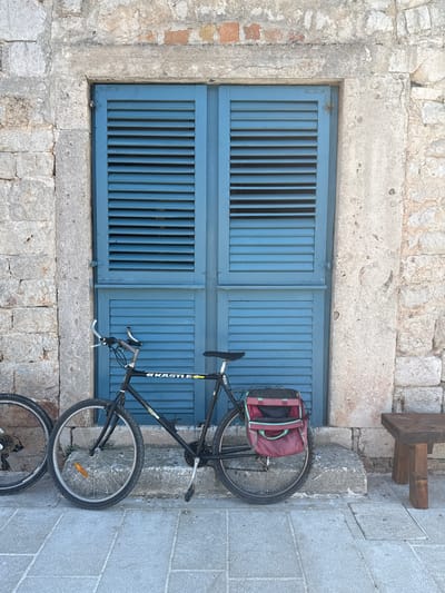 A bicycle with a red bag attached to the rear rack leans against a weathered stone wall in front of blue louvered shutters. The bicycle is black with silver handlebars and appears to be a mountain bike. Part of another bicycle's wheel is visible to the left of the main bicycle. The stone wall is constructed from rough-hewn blocks and shows signs of age and weathering. The shutters are a vibrant blue and are slightly ajar. A small, rustic wooden stool or bench is positioned to the right of the bicycle. The ground in front of the scene is paved with grey stone tiles. The overall impression is of a rustic, possibly Mediterranean, setting, consistent with a location like Jelsa, Croatia, known for its stone architecture and charming streets.