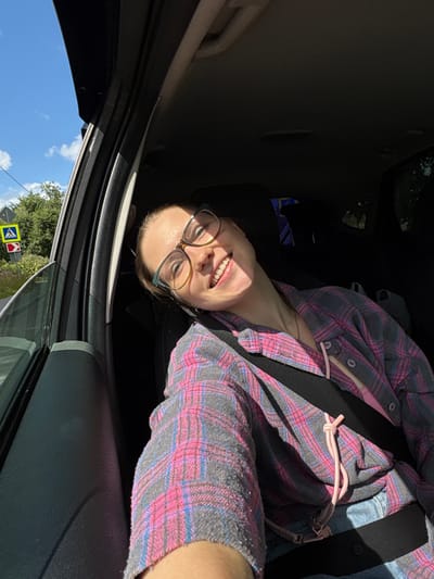 A young woman with glasses is sitting in a car, her head tilted back and a wide smile on her face, looking directly at the camera. She is wearing a pink and gray plaid shirt and a seatbelt is across her chest. The car interior is dark, but sunlight streams in from the open window, illuminating her face. Outside the window, a blue sky with white clouds is visible, along with green trees and a road sign with yellow and red markings. The scene appears to be captured during the daytime in a semi-urban or rural setting, potentially in the city of Imeni Sverdlova, Russia, given the presence of trees and the style of road signage.