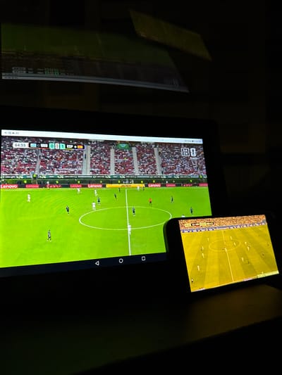 The image depicts a dark room where two electronic screens are displaying a live football match. The larger screen, likely a tablet, shows a close-up view of the game with score graphics indicating "URU 0 1 ESP" and the time as "64:55". The field is green, with players in white and dark blue uniforms visible. The stands behind the players are filled with spectators. The smaller screen, a smartphone, is angled to the right and also displays the football match, though with a wider view of the field and different advertising on the sidelines. The overall environment is dimly lit, with the glow of the screens being the primary light source. The presence of the "Lenovo" logo on the larger screen suggests it is a Lenovo device. The image has been captured from a slightly elevated viewpoint looking down onto the screens. The context of Seixal, Portugal is not evident from the visual information provided in the image itself.