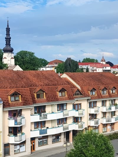 The image captures an aerial view of a residential building with a reddish-brown tiled roof and cream-colored facade. The building features multiple balconies, many of which are adorned with railings and some with plants. The lower levels of the building appear to house commercial establishments, indicated by storefronts and signage.
In the background, a church tower with a dark spire and a clock face stands prominently against a cloudy sky. The surrounding urban landscape includes other buildings with red roofs, trees, and a distant church dome. The presence of the church tower and the architecture of the buildings strongly suggest the location is in Nový Jičín, Czechia. The scene depicts a typical urban environment with residential and commercial structures, a church, and foliage.