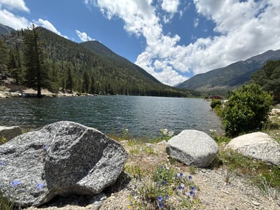 This image captures a serene natural landscape, likely taken during the daytime with sunny weather and scattered clouds. The scene features a tranquil lake in the foreground, reflecting the bright sky and surrounding mountains. The water appears calm with gentle ripples.
The shoreline is dotted with large, grey granite boulders, some covered in sparse green vegetation and small purple and white wildflowers. A narrow path or trail is visible along the edge of the water, made of gravel and small stones.
In the midground, a dense forest of evergreen trees covers the slopes of a majestic mountain. The trees are a vibrant green, creating a lush texture on the mountainside. Further in the distance, more mountains rise, some appearing rocky and less vegetated, hinting at a higher elevation.
On the right side of the lake, a small, red building with a white roof can be seen nestled amongst the trees. This structure suggests a human presence in the otherwise wild landscape, possibly a cabin or a small visitor center. A few scattered trees and bushes are also visible on this side, contrasting with the dense forest on the left.
The overall mood of the image is one of peace and natural beauty. The composition, with the expansive lake and towering mountains, evokes a sense of awe and tranquility. The clear blue sky with fluffy white clouds adds to the picturesque quality of the scene. The location is identified as Nathrop, United States, suggesting a mountainous, perhaps alpine, environment. There is no visible text in the image.