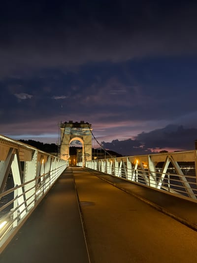 The image captures a low-angle, wide-shot view of the Pont du Rose, a suspension bridge in Vienne, France, at dusk. The sky is a dramatic blend of deep blues, purples, and pinks, with dark, textured clouds indicating the end of daylight. The bridge itself is made of white painted metal, with a sturdy railing running along its length. Lights illuminate the bridge's structure, casting a warm glow on the walkway, which appears to be asphalt. The bridge leads towards a stone archway, part of a larger structure, possibly a bridge tower, which is illuminated and shows some graffiti and scaffolding. Beyond the archway, distant buildings and a tree-covered hillside are visible against the twilight sky. The overall scene is tranquil and atmospheric, highlighting the architectural features of the bridge against the backdrop of a colorful sunset.
