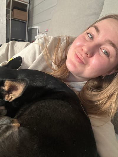 A young woman with blonde hair and light eyes is taking a selfie. She is wearing a light-colored hoodie. Part of a black dog with tan ears is visible to her right. The background shows a room with white walls, a window, and some objects on a shelf. There is no indication of Bashur, Russia in the image.