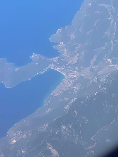 This is an aerial view of a coastal area, likely taken from a high altitude given the curvature of the Earth visible on the right side of the frame. The image showcases a stretch of land meeting a body of blue water. The landmass appears to be heavily textured with various shades of white and gray, suggesting a mix of terrain, possibly mountains or hills, with some lighter patches that could indicate settlements or vegetation. The coastline is irregular and rugged, with peninsulas and inlets. The water is a deep, clear blue. The lighting suggests it is daytime, and there are no obvious indications of specific weather conditions, though the slight haziness could imply a bit of atmospheric moisture or distance. The overall scene depicts a natural landscape with hints of human presence through what appear to be scattered structures or roads visible on the land. Given the location context of Menteşe, Turkey, this could be a view of the Aegean or Mediterranean coastline, characterized by its mountainous terrain and beautiful blue waters. There is no visible text in the image.