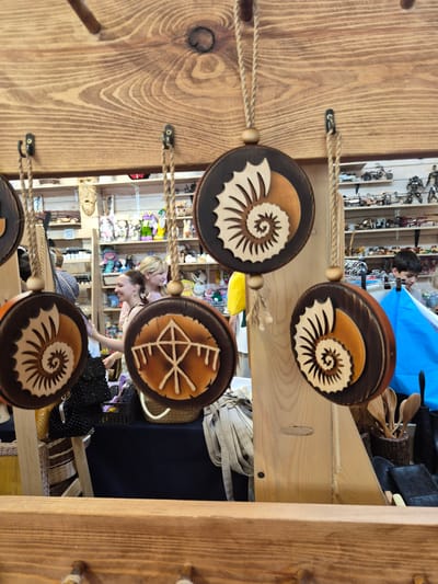 The image depicts an indoor market stall with wooden shelves displaying various items. In the foreground, several circular wooden ornaments with carved ammonite designs are hanging from ropes. Behind the ornaments, a man and two women are visible. The women appear to be browsing the stall, and one is interacting with a display of goods. The shelves behind them are filled with a variety of objects, including what appear to be small toys and decorative items. The overall impression is of a lively marketplace. The city of Krasnyy Klyuch, Russia, is mentioned as the location, suggesting a cultural context for the market and its wares.