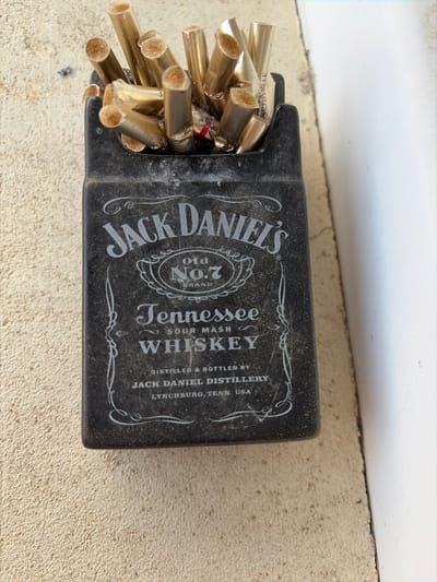 The image shows a black cigarette pack designed to resemble a bottle of Jack Daniel's Tennessee Whiskey. The pack is full of gold-filtered cigarettes. The pack and cigarettes are resting on a light beige surface, possibly concrete, next to a white wall with a painted edge. No people are visible. The image does not contain information that allows location in the city of Kubrat, Bulgaria, to be inferred.
