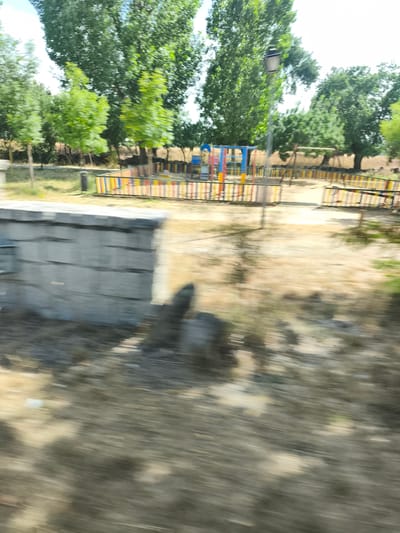 The image depicts a blurred outdoor scene, likely a park or playground, with a motion blur suggesting movement. In the background, trees with green foliage dominate the landscape. A brightly colored playground structure, with elements of blue, yellow, and red, is visible behind a fence. The fence itself is composed of colorful vertical slats in shades of yellow, orange, blue, and red. A lamppost stands prominently near the playground. In the foreground, there is a low, gray, stone-like wall, possibly part of a planter or boundary. The ground appears to be covered in dry, light-colored earth or sand, with patches of green vegetation. The overall impression is of a sunny day, given the bright lighting and the vibrant colors. The motion blur creates a sense of dynamism and speed.