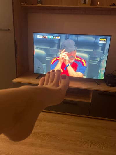 In the foreground of the image, a bare foot with dark red nail polish is visible, positioned on a light brown wooden floor. The foot is angled towards a television screen that is displaying a soccer match. The television screen shows a man wearing a dark baseball cap and a red, yellow, and blue soccer jersey, appearing to be a coach or manager. A scoreboard overlay on the screen indicates the time as 47:52, with the score of POR 0 - 0 ESP. In the upper right corner of the screen, a logo reads "МАТЧ!" and "ПРЯМОЙ ЭФИР". The television is placed on a dark brown and gray entertainment unit. The background shows a light brown wooden shelf mounted on a wall. The overall scene appears to be an indoor living space, likely a home, in the city of Lesnye Polyany, Russia.