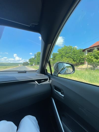 The image is a first-person view from inside a vehicle traveling on a road. The interior of the car is visible in the foreground, with the dashboard, door panel, and a portion of the passenger's leg covered in white fabric. The passenger side window offers a view of the exterior.
Outside the car, a rural landscape unfolds. A paved road stretches into the distance, bordered by green grass and trees. On the right side of the road, a house with a red tile roof is partially visible, surrounded by vegetation and a fence. The sky is blue with scattered white clouds. The scene appears to be set in Coșernița, Moldova, on a bright, sunny day.